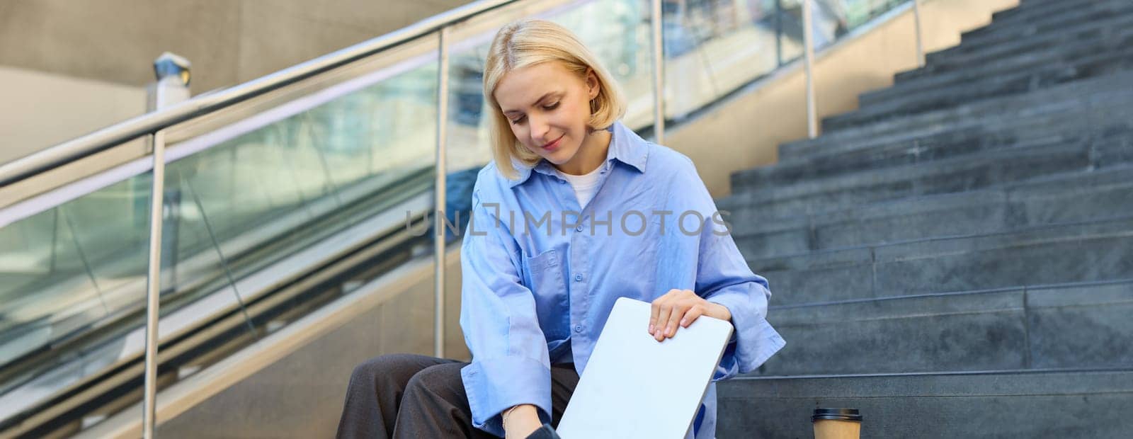 Portrait of young street-style girl, student with backpack, putting away laptop, packing, sitting on stairs with cup of coffee, wearing blue shirt.