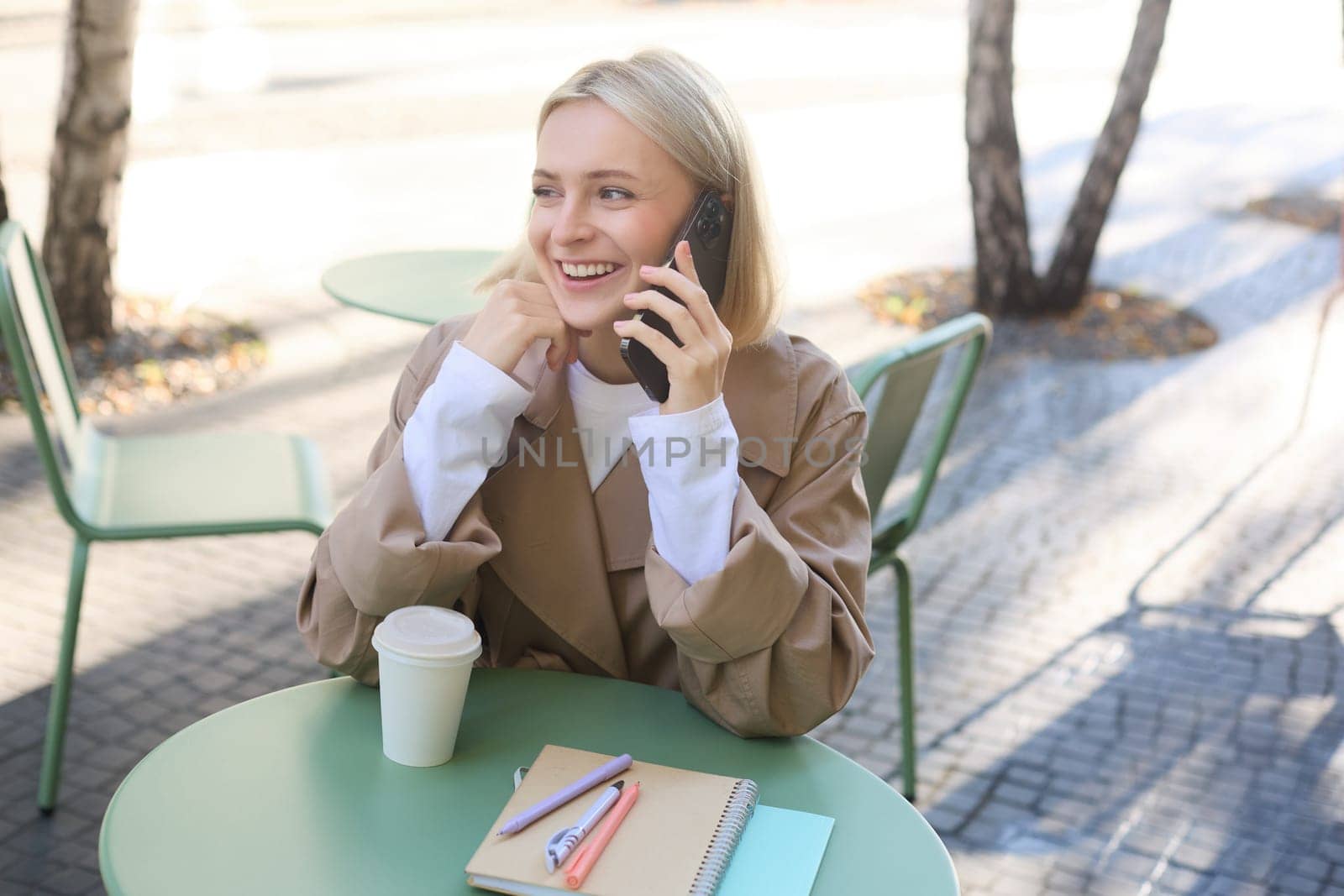 Carefree young woman in trench, talking on mobile phone and smiling, sitting outside cafe and drinking coffee, answer a call.