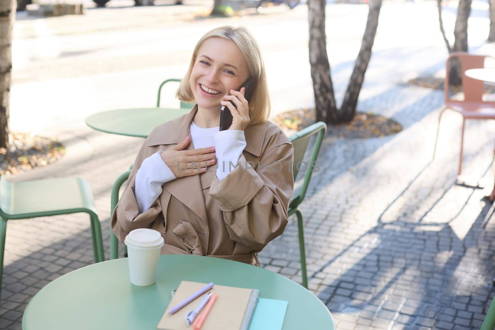 Image of stylish blond woman talking on mobile phone, drinking coffee in cafe outdoors, enjoying warm weather in city centre, answer telephone call on smartphone by Benzoix