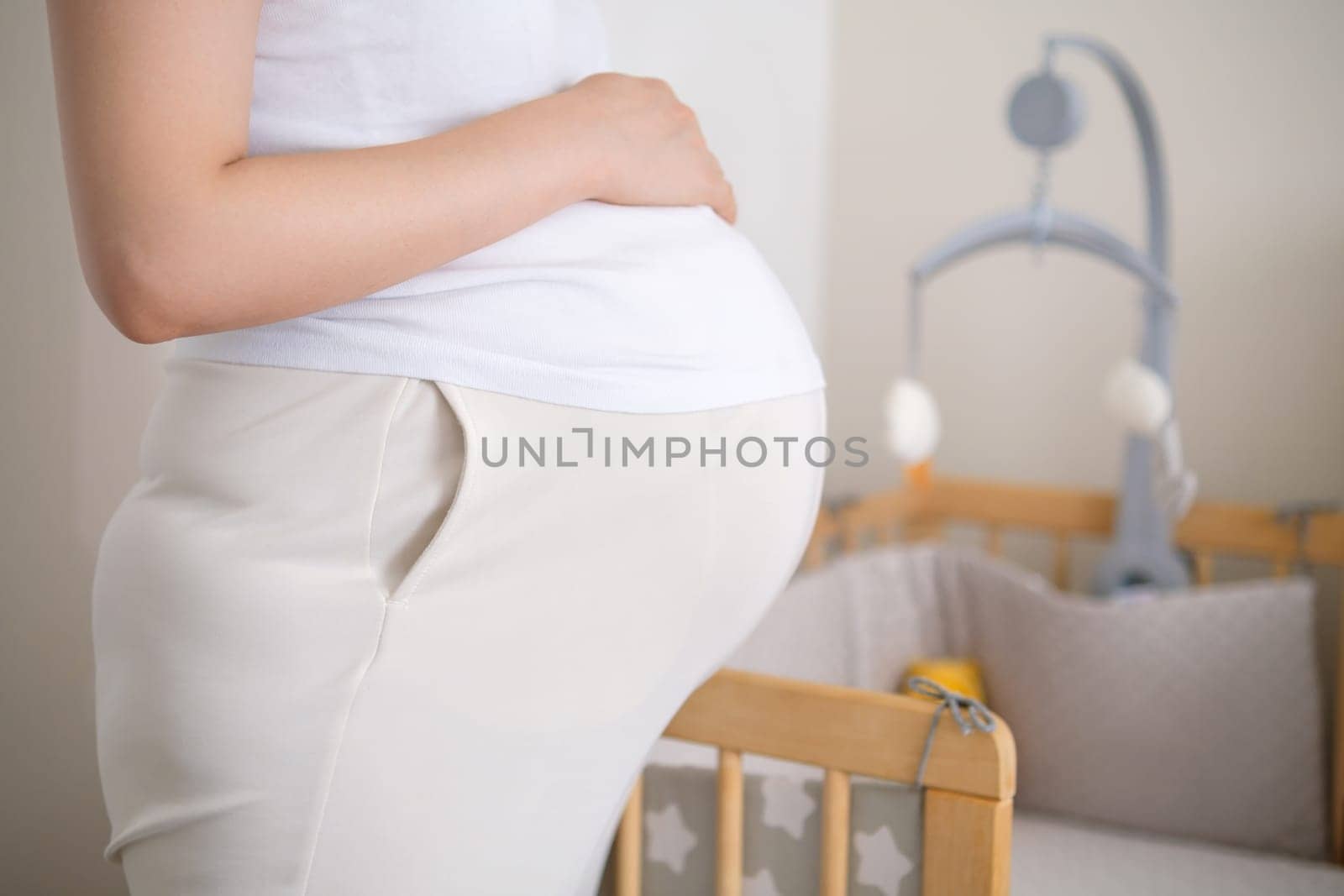 Pregnant woman stands near baby bed with hanging toys. Woman fingers trace contours of crib simultaneously caressing belly in anticipation of baby