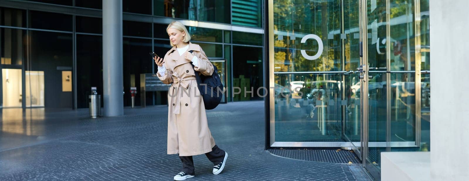 Portrait of blond young woman walking on street, student with backpack looking at her mobile phone, reading message on smartphone by Benzoix