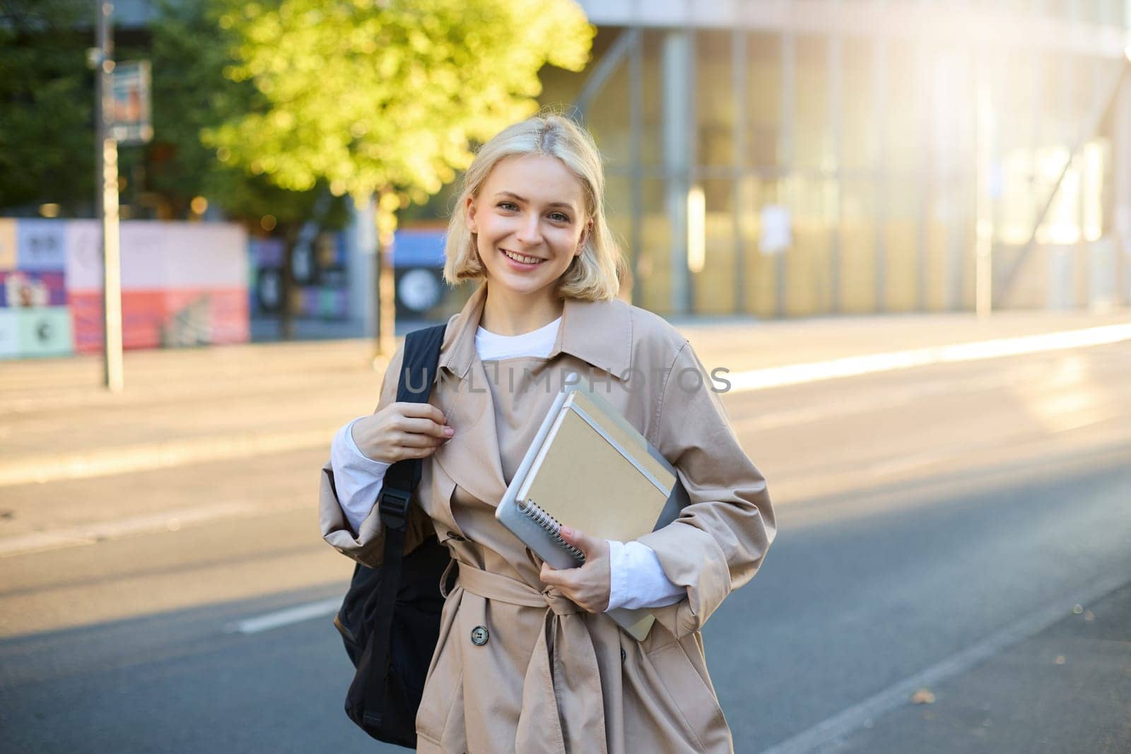 Portrait of young stylish female student, woman with backpack and notebook, smiling happy at camera, standing on street on sunny autumn day.