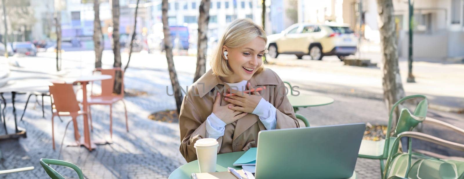 Lifestyle and technology concept. Young blond working woman in trench coat, studying, working on project on laptop, sitting in coffee shop on sunny day, making notes, attending online course by Benzoix