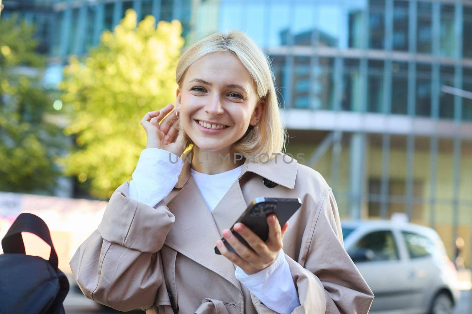 Portrait of carefree smiling woman on street, holding mobile phone, tuck hair behind ear and looking happy at camera, waiting for someone outside.
