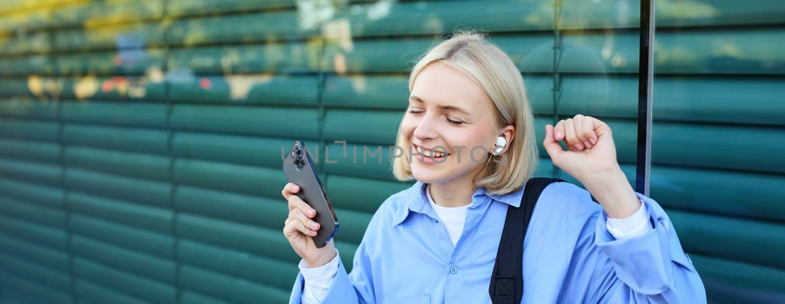 Close up portrait of blond smiling woman, wearing shirt, holding backpack and mobile phone, dancing and listening to music in wireless headphones, closes eyes and raises hand up.