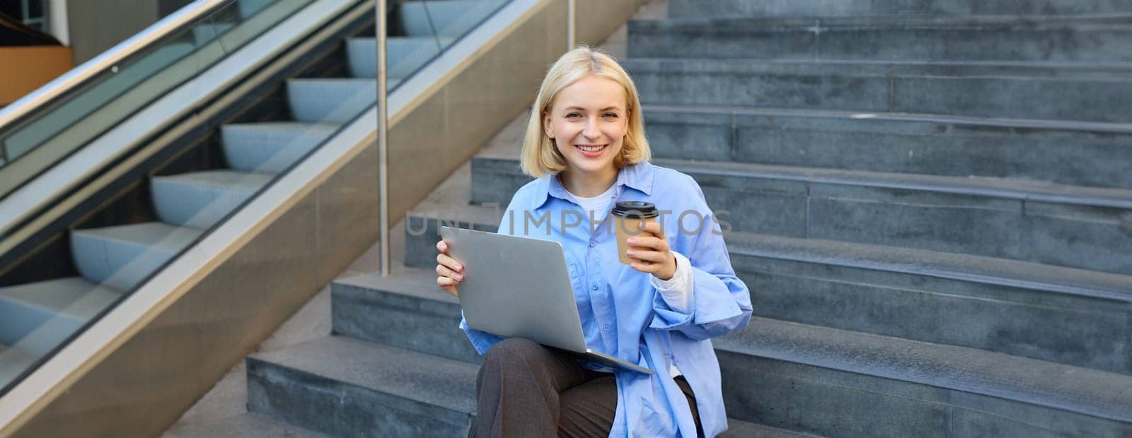 Young woman sitting with cup of coffee and laptop on stairs outdoors, working remotely, freelancing, connecting to public wifi in city to study online by Benzoix