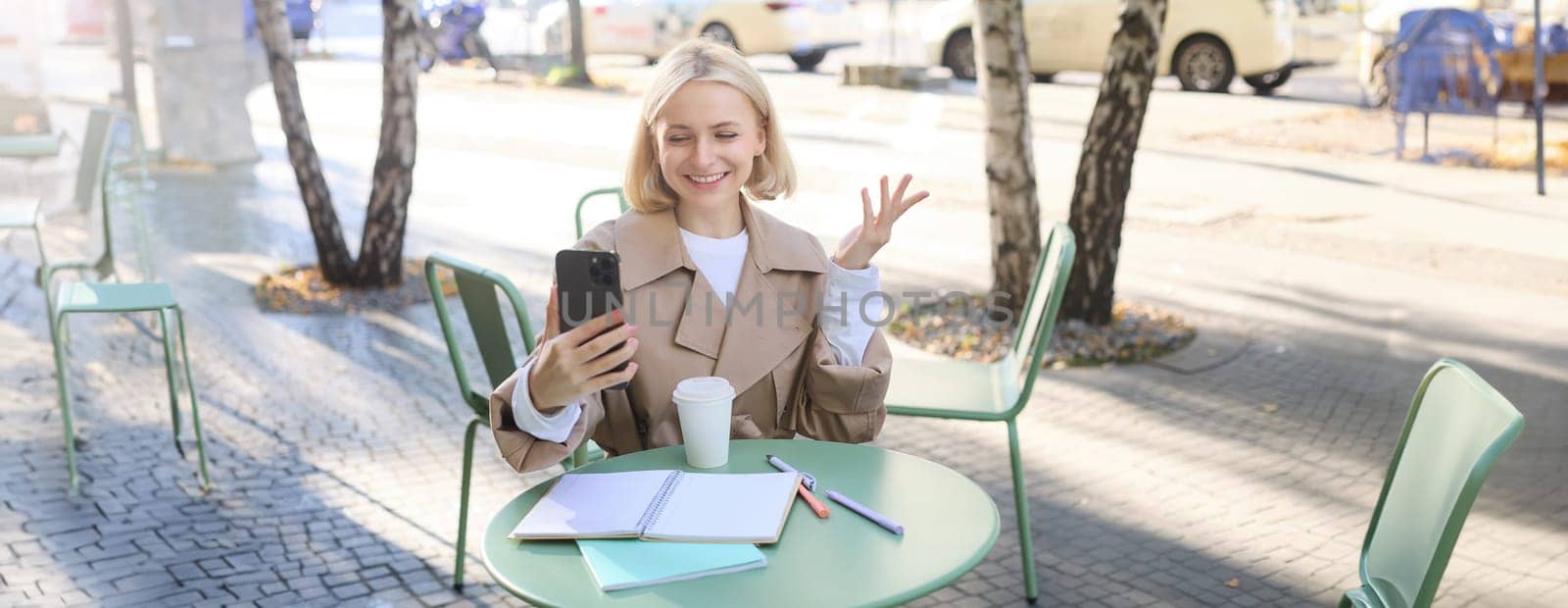 Image of young woman working outdoors in cafe, sitting outside with smartphone, video chatting, connecting to online meeting using mobile phone, smiling and looking happy.