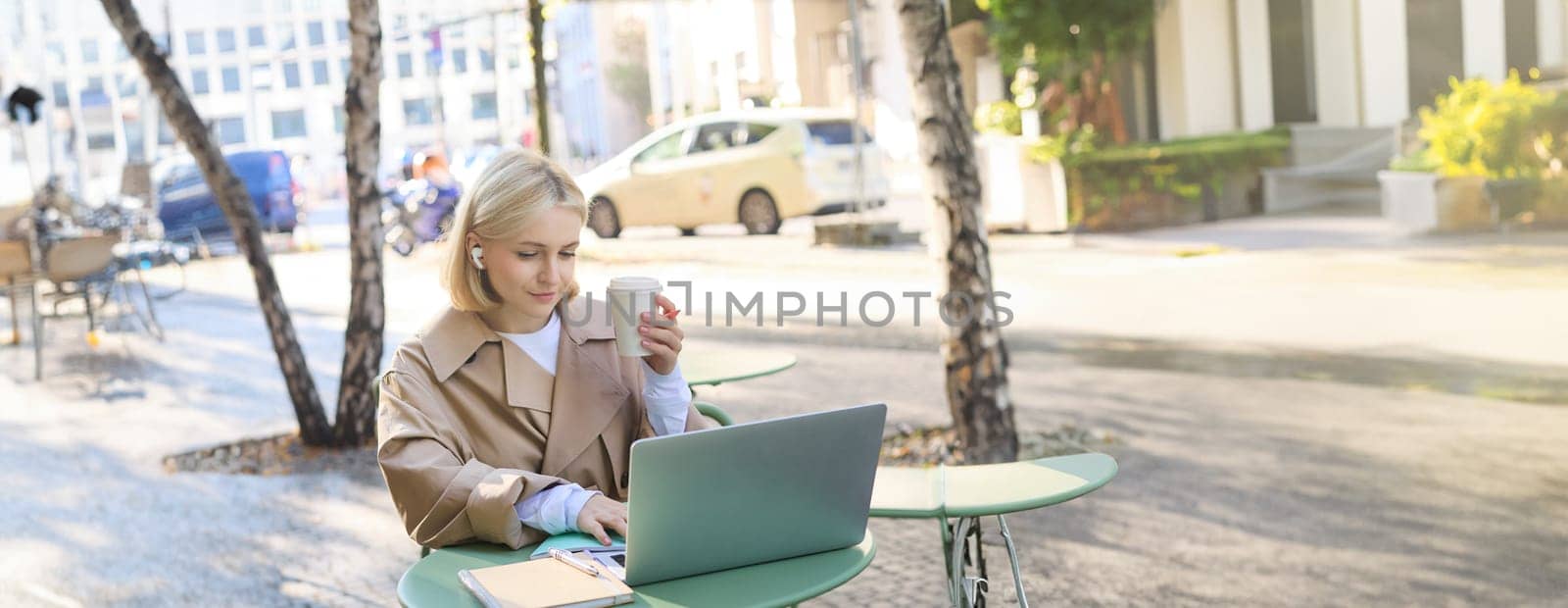 Vertical shot of young blond woman with laptop, sitting with coffee in street cafe, working outdoors on computer, wearing wireless headphones and smiling.