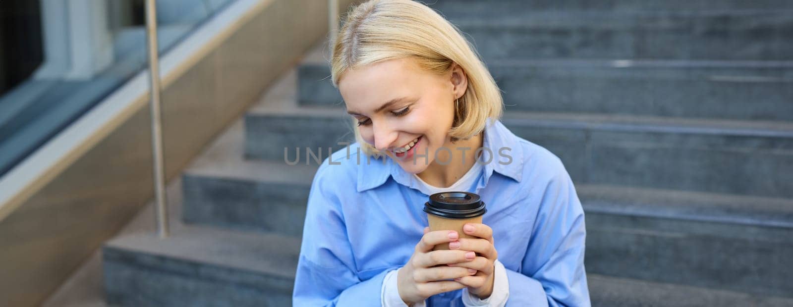 Close up portrait of beautiful, smiling blonde woman, student sitting on stairs outside campus, drinking takeaway coffee, warm-up her hands while holding a cup.