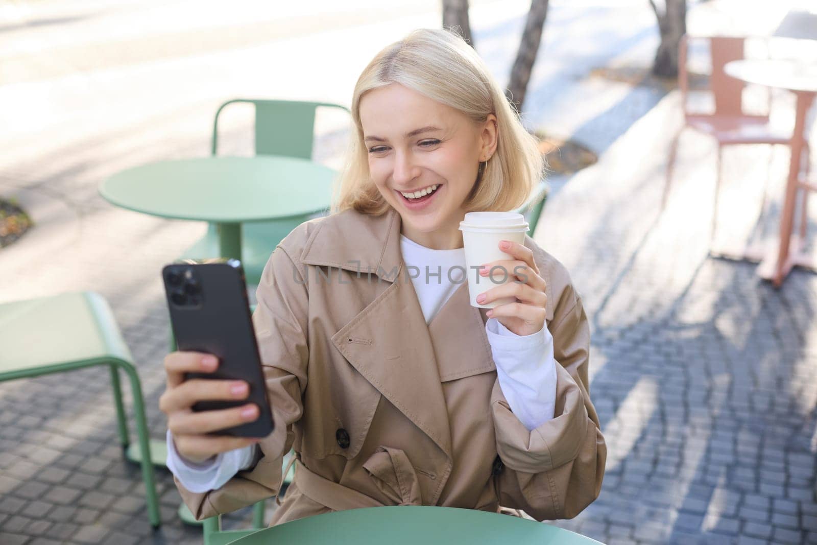 Stylish young woman, social media influencer, taking selfie, video chatting, using smartphone, drinking coffee outdoors in cafe.