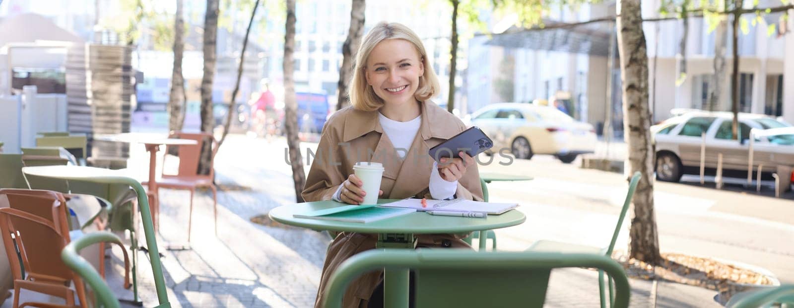 Portrait of modern young woman in city centre, sitting in cafe outdoors, drinking coffee, using mobile phone, smiling.