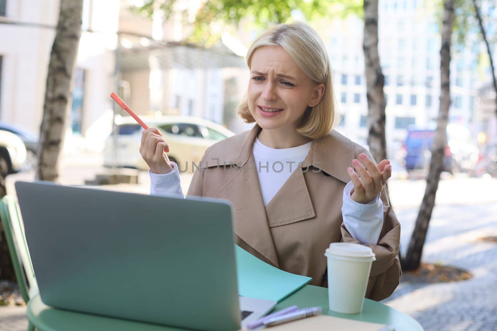 Portrait of young female student, modern woman in outdoor coffee shop, street cafe, talking to someone via laptop, video chatting, communicating, working remotely online by Benzoix