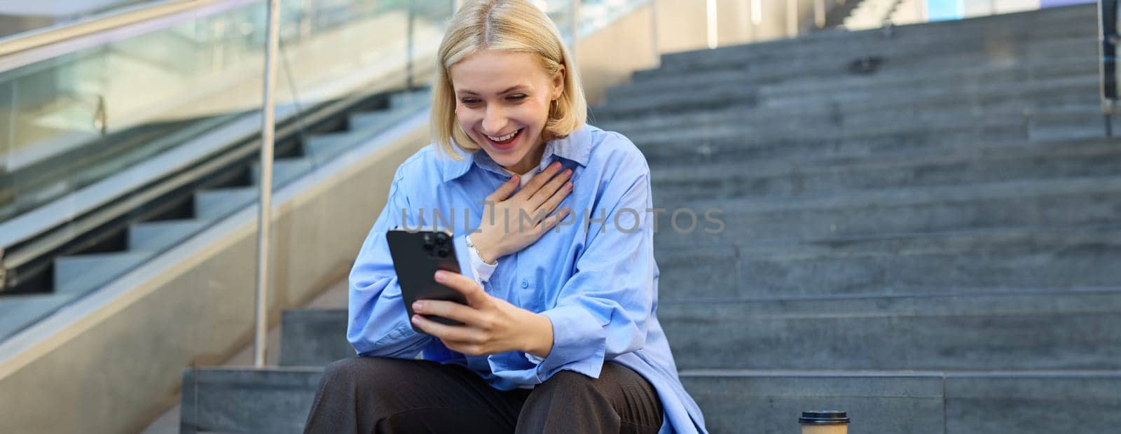 Portrait of young smiling woman, looking at smartphone with pleased, happy face, holding hand on chest, pleased to see something, sitting on stairs outdoors.