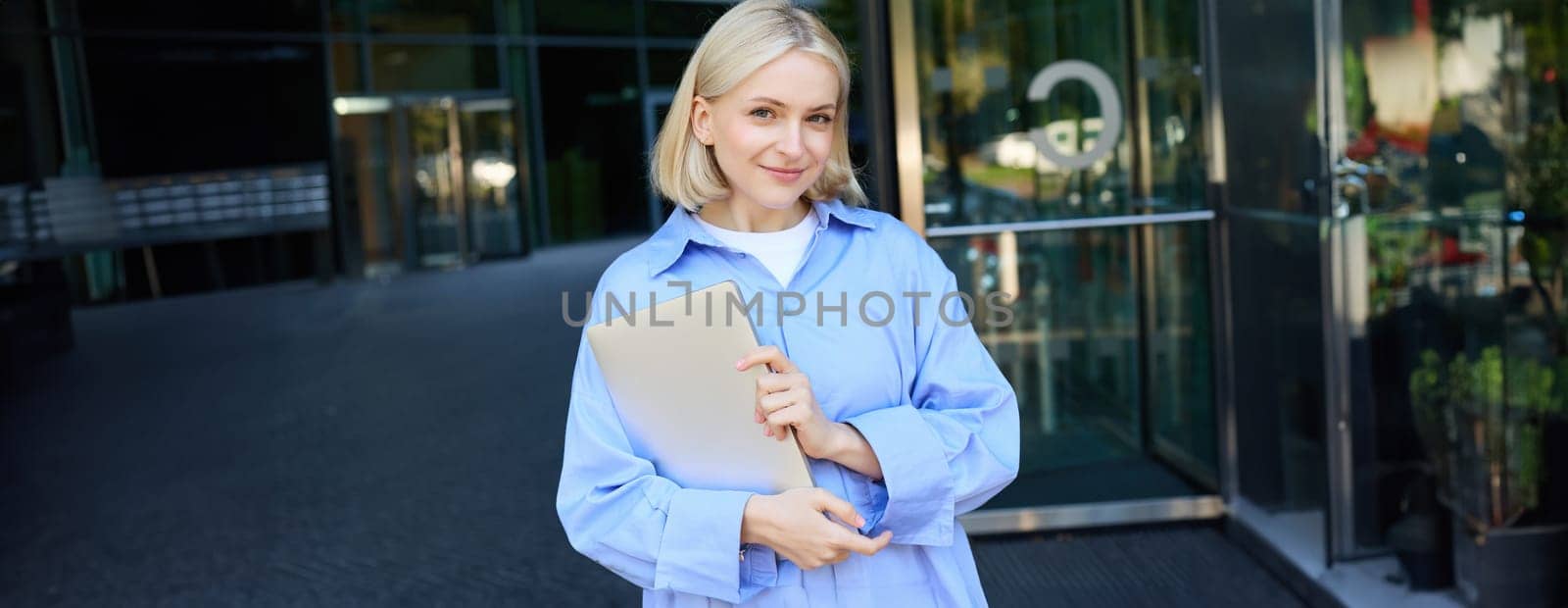 Image of young female employee, working woman standing near her office on street, holding pile of documents and smiling by Benzoix