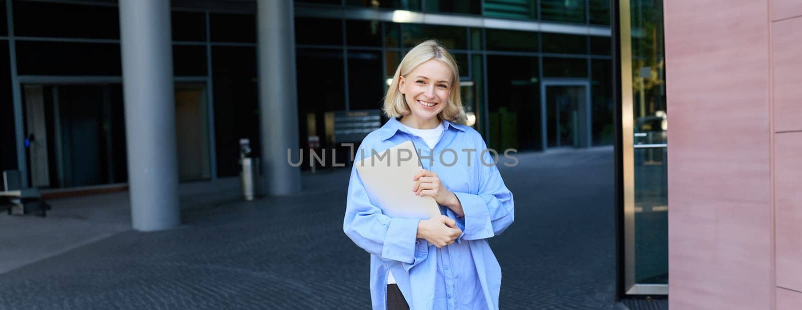 Portrait of young blond woman, student standing near her campus with notebooks and documents, wearing blue shirt and smiling at camera. Education concept