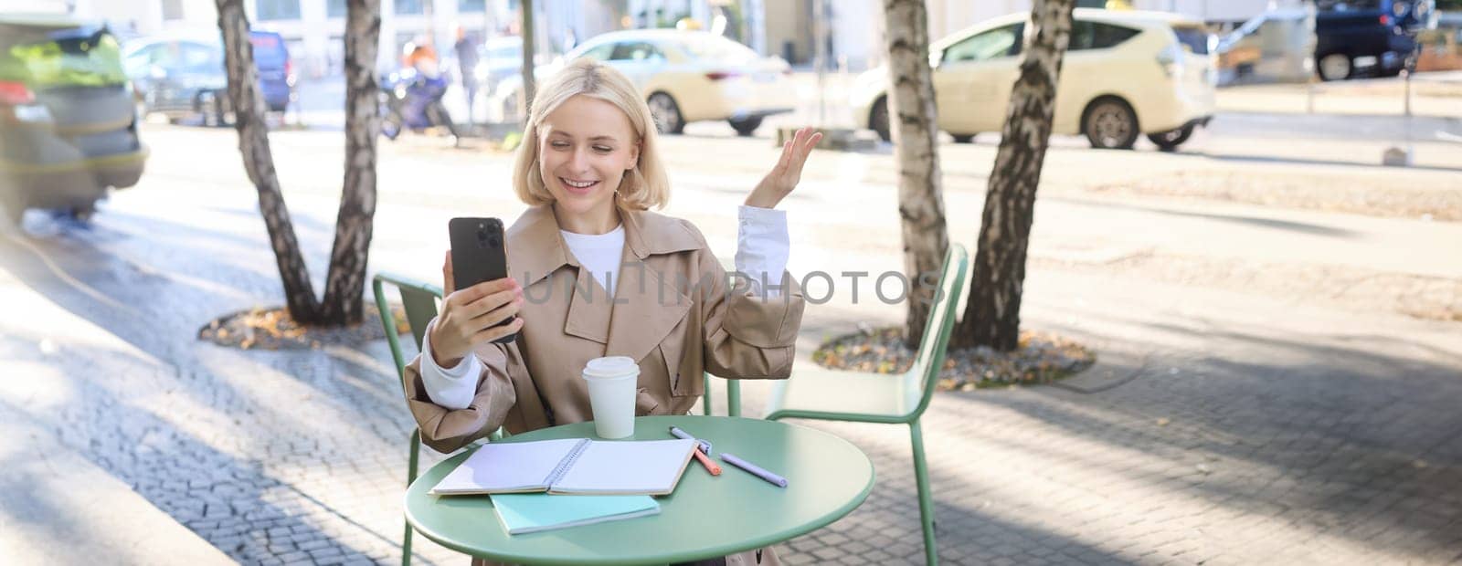 Portrait of young stylish woman in trench, sitting outdoors in city cafe, talking on video chat, using mobile phone to connect to online conversation, drinking coffee by Benzoix