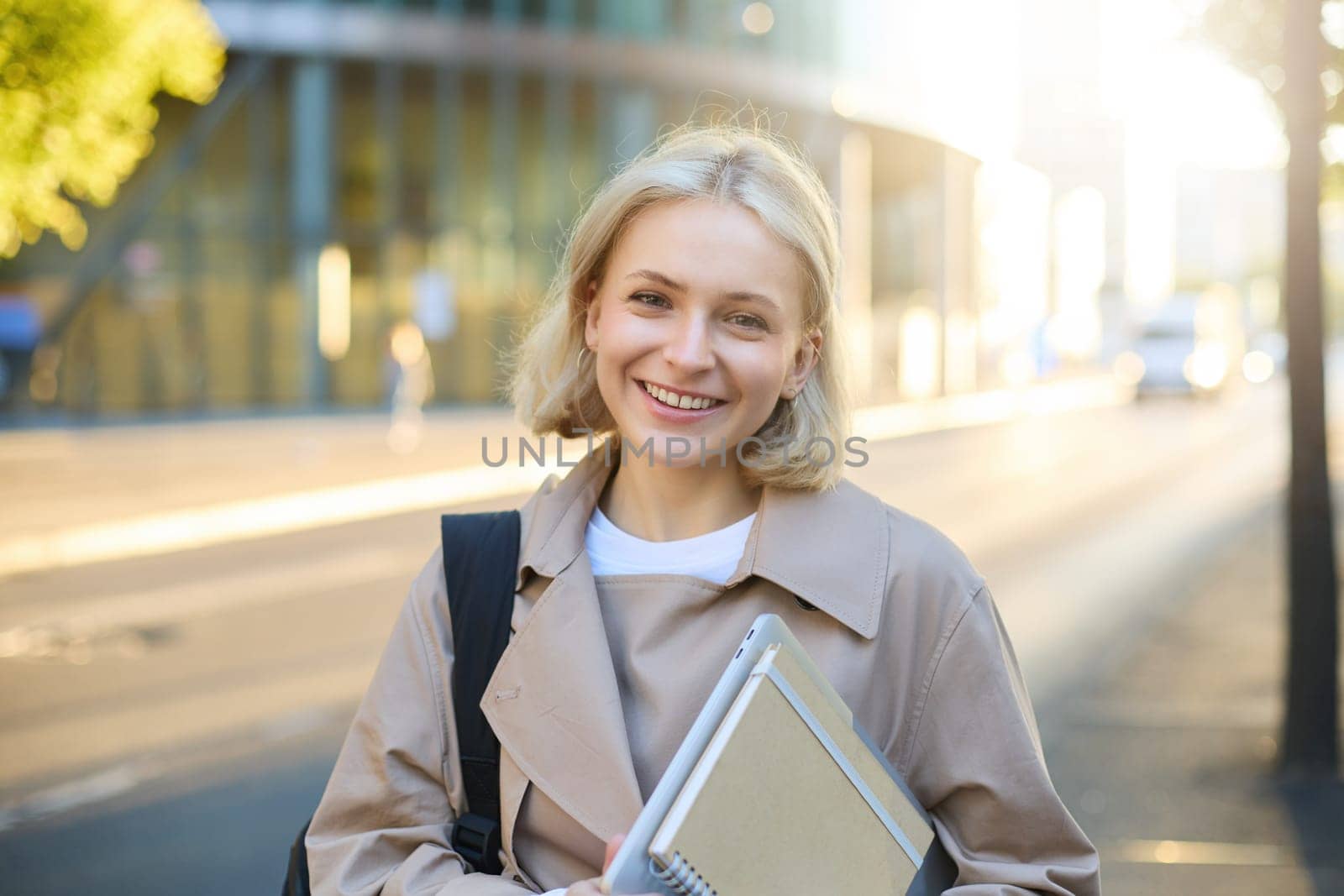 Image of beautiful young smiling woman with blond hair, walking along street on sunny day, carry her notebooks and college materials, looking carefree by Benzoix