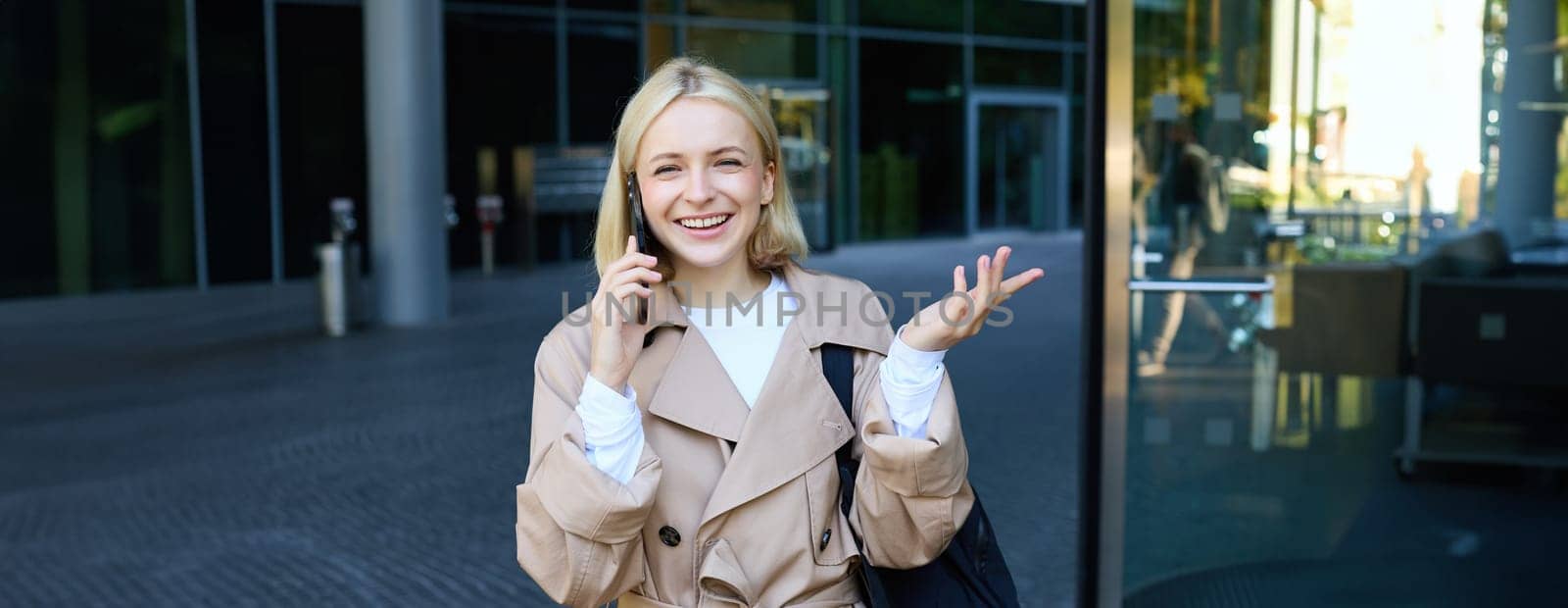 Carefree young woman with smartphone, walking outdoors in city centre, standing on street and chatting, talking on mobile phone with happy, cheerful smiling face expression by Benzoix