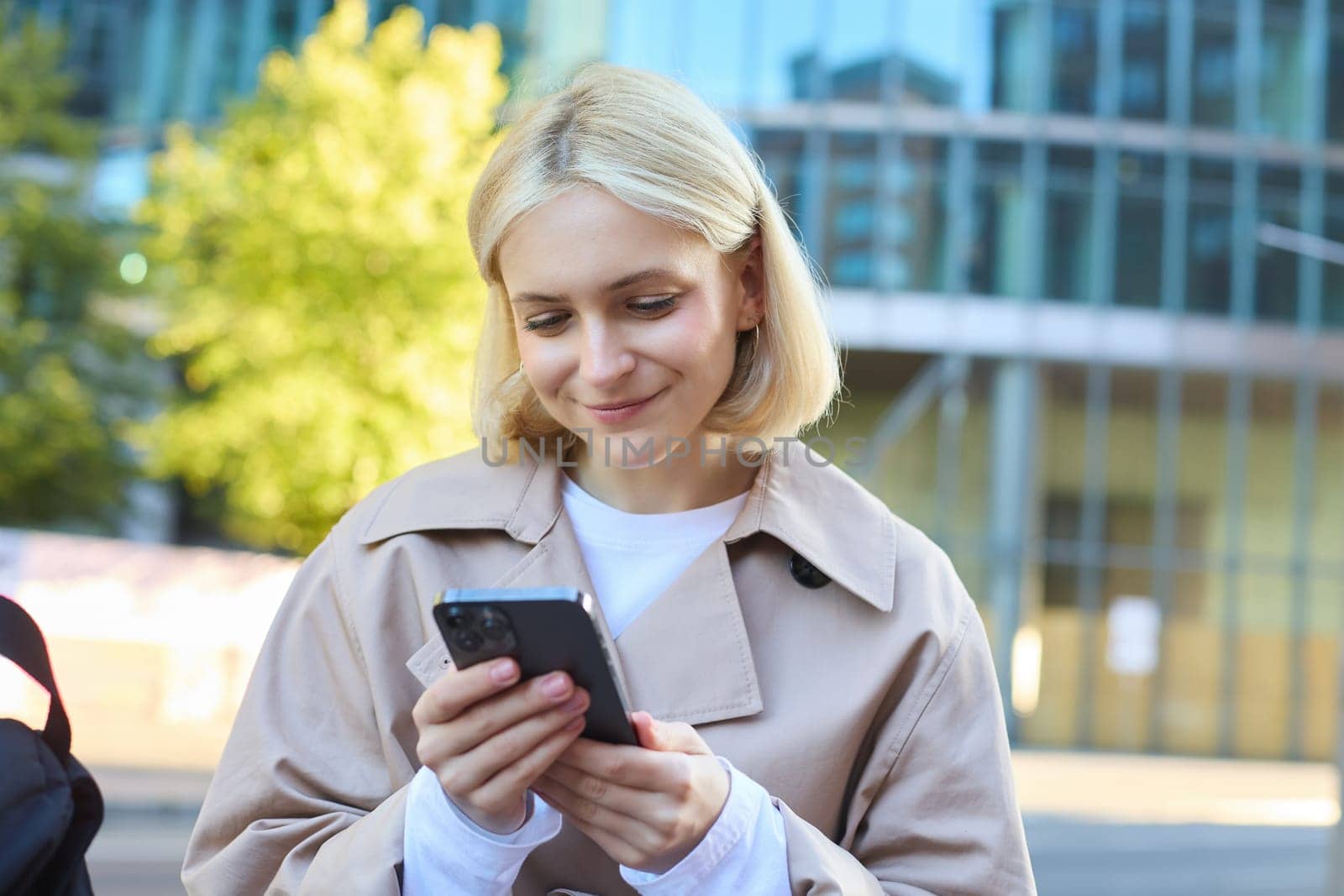 Close up portrait of smiling female model on street, sitting on bench with mobile phone, looking at smartphone screen, messaging with friend while waiting outside.