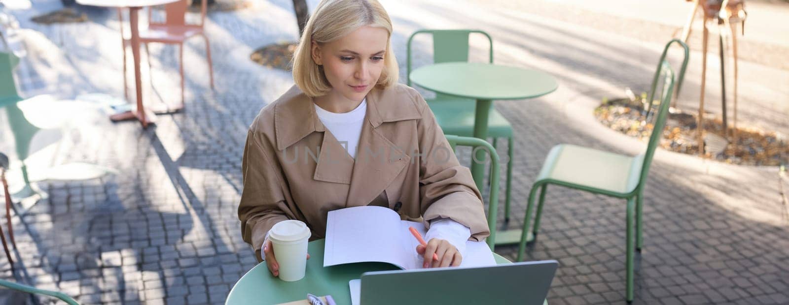 Image of young woman studying outdoors, sitting in cafe, making notes while listening to online lecture using laptop, making notes in notebook, writing by Benzoix