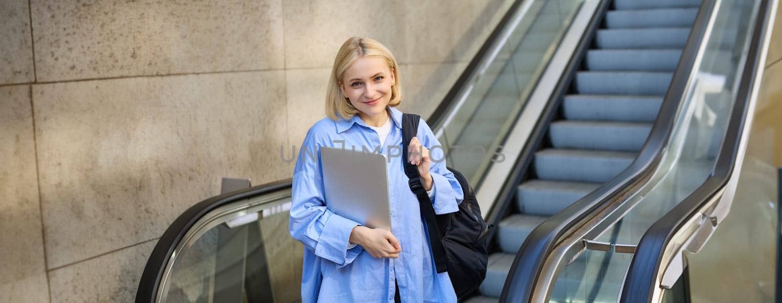 Education and people concept. Young woman with laptop and backpack, using escalator in city centre, smiling and looking at camera.