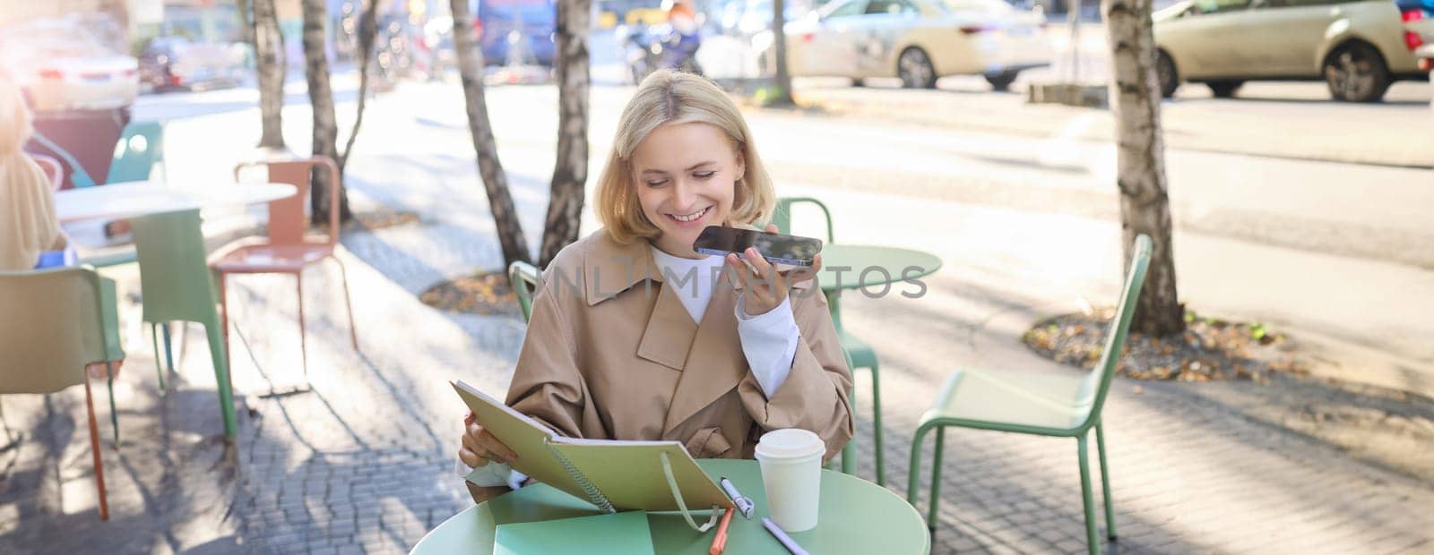 Lifestyle portrait of happy young woman, sitting in cafe and recording voice message, holding mobile phone near lips and talking into microphone, spending time in cafe, drinking coffee and chatting by Benzoix
