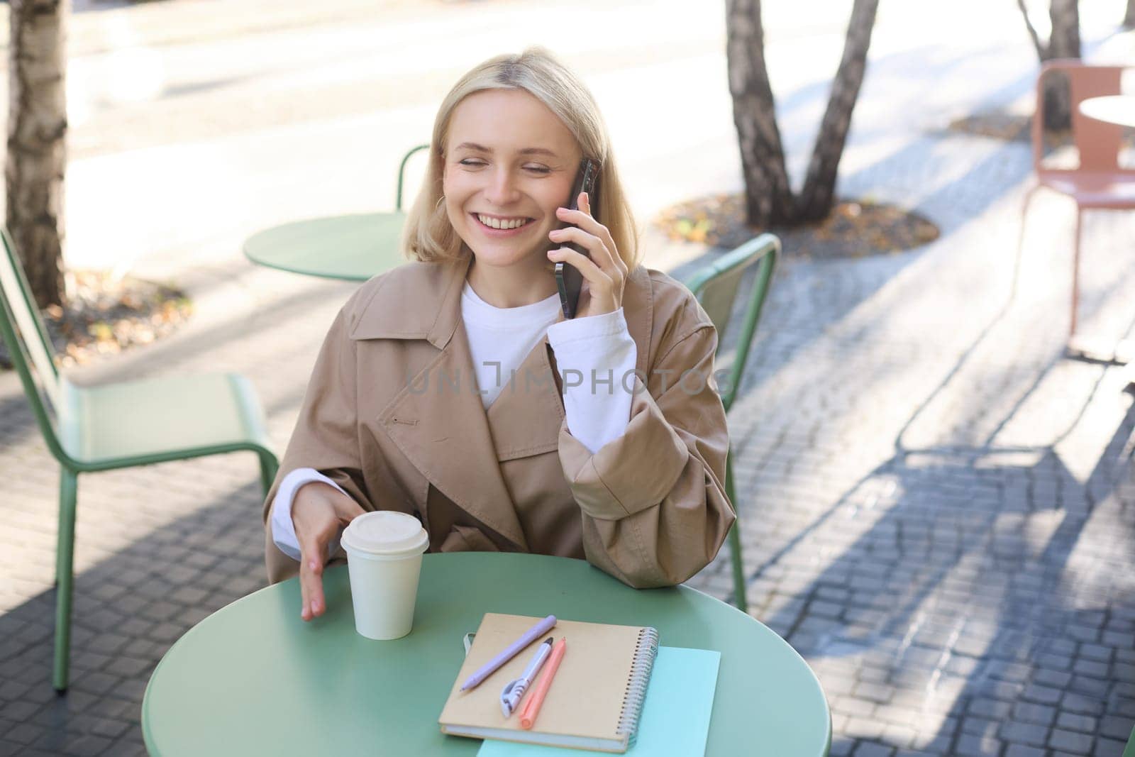 Cheerful young woman laughing, smiling while talking on mobile phone, sitting in cafe outdoors, enjoying friendly conversation.