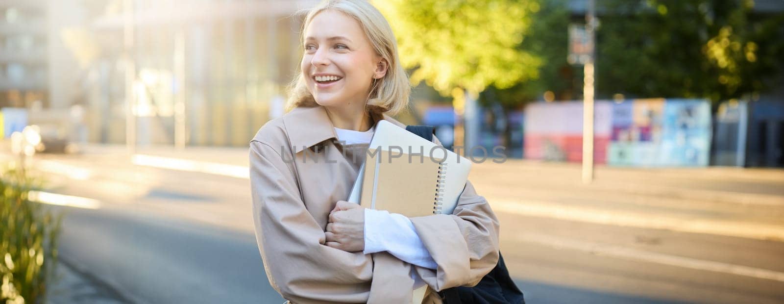 Candid young woman on her way to University, standing on street, carries notebooks and homework, has backpack on shoulder, turns away, laughs and smiles, shows genuine emotions.
