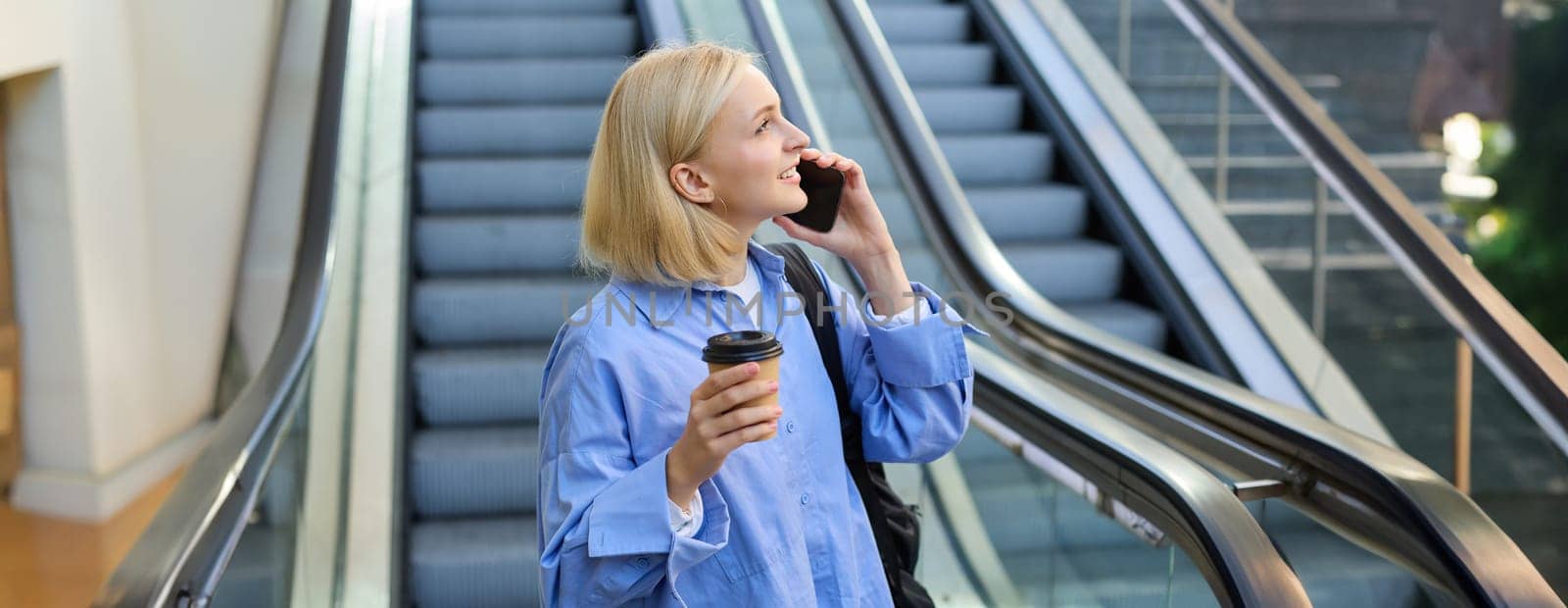 Image of young stylish woman, girl with cup of coffee, waiting for friend near escalator, talking on mobile phone, answers a call, looking up.