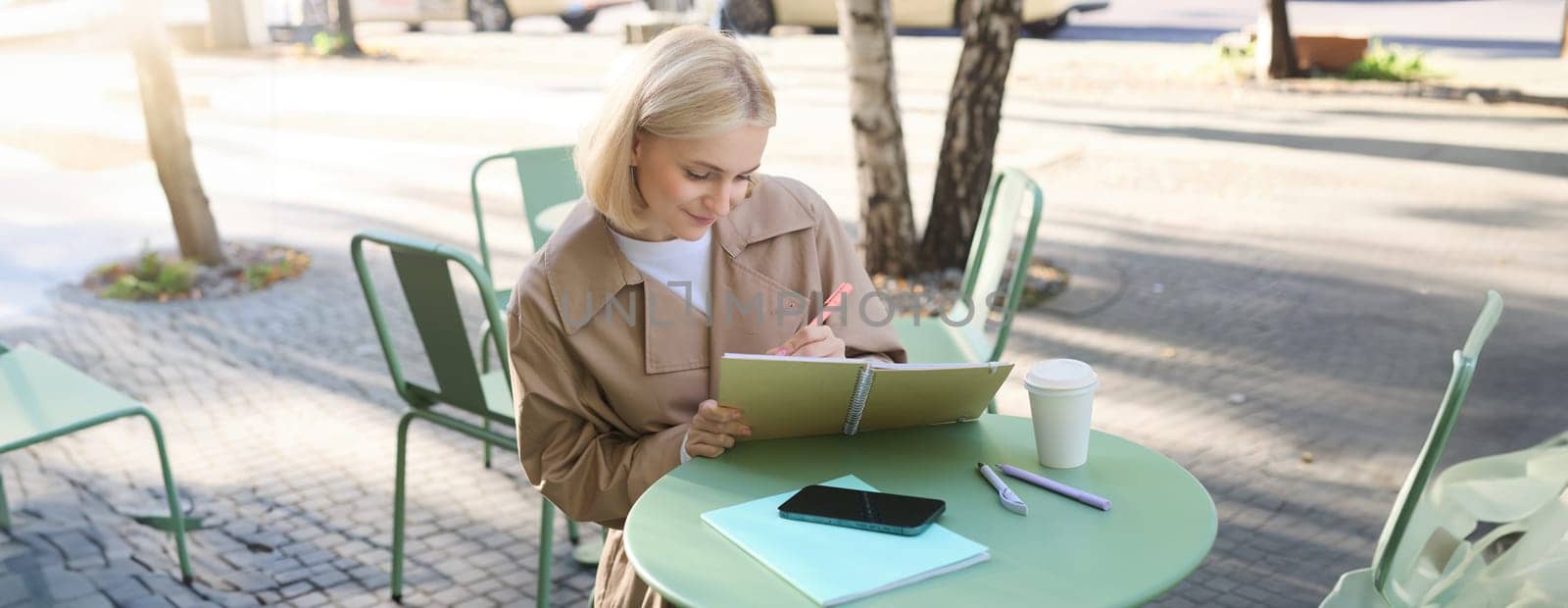 Portrait of beautiful blond woman, sitting in outdoor coffee shop, drawing in cafe in notebook, making sketches outside on street,