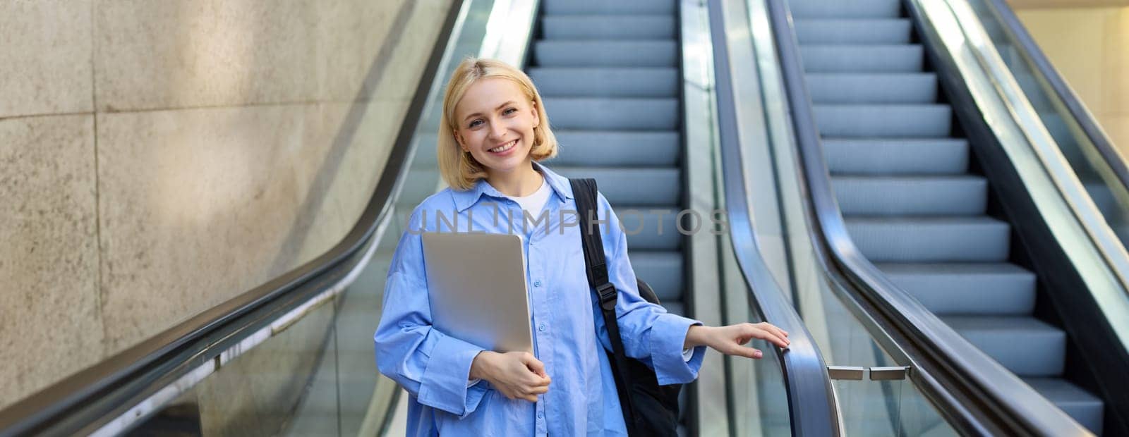 Portrait of smiling, beautiful blond girl, student with laptop and backpack, wearing blue shirt, using escalator, going down, looking confident at camera.