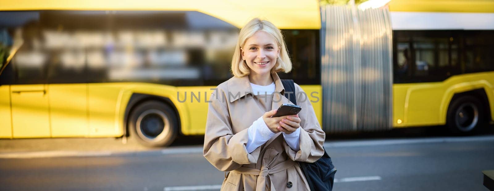 Outdoor shot of young woman with backpack, standing on street with cars passing behind her, holding mobile phone and smiling, urban lifestyle concept.