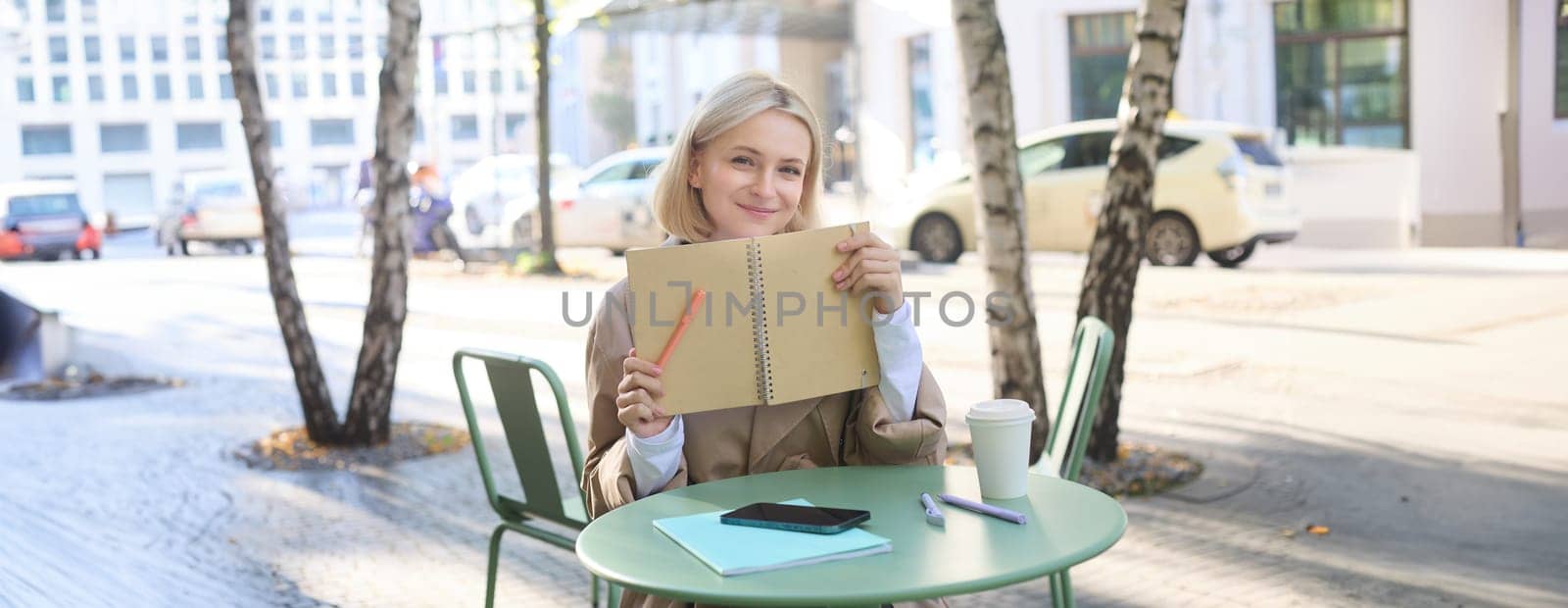 Image of young modern woman, student sitting in coffee shop with smartphone, holding notebook, showing her yellow journal, writing in planner and smiling by Benzoix