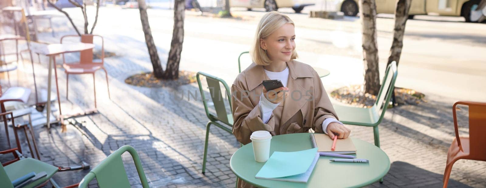 Image of young beautiful girl, student studying, doing homework in cafe, using mobile phone, holding smartphone, smiling.