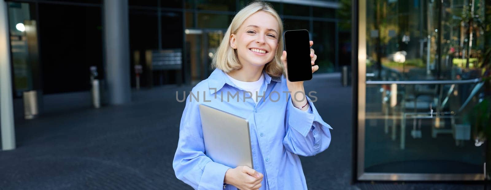 Happy smiling woman showing smartphone screen, demonstrating mobile phone display, standing in college outdoors, posing near campus with laptop and telephone by Benzoix