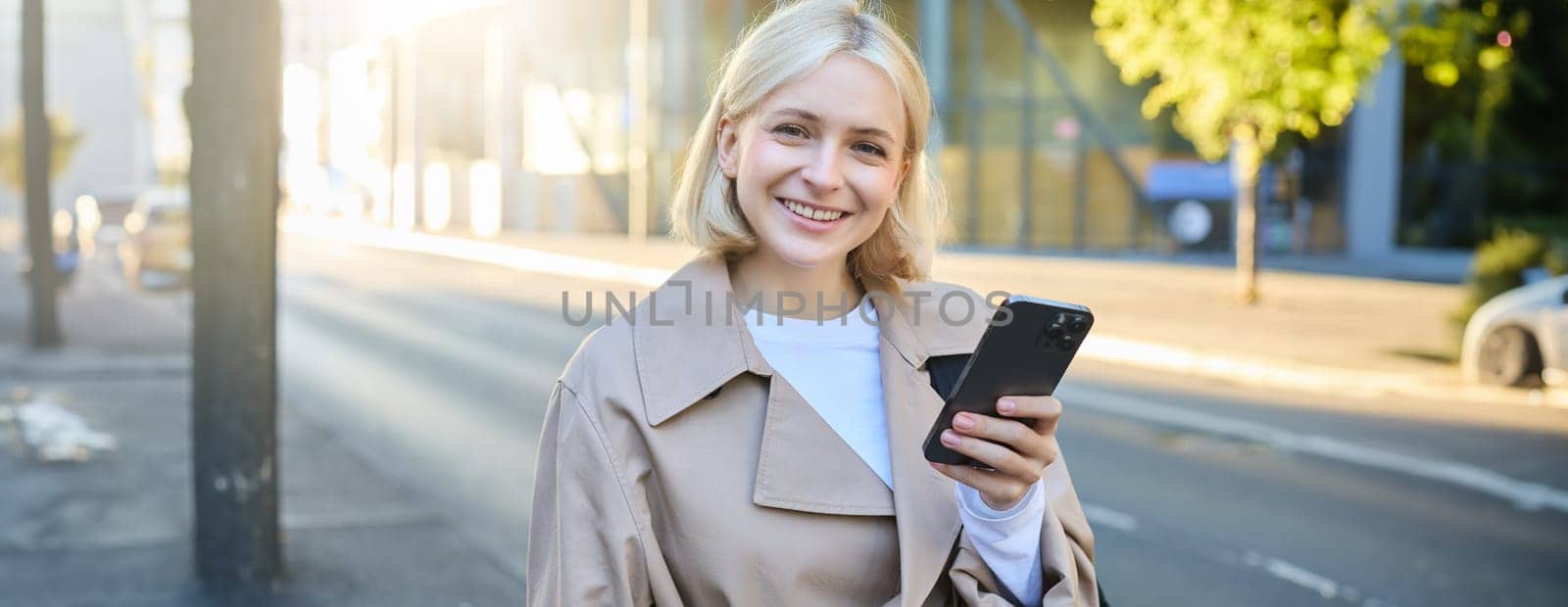 Portrait of smiling blonde woman, wearing trench coat on street, looking happy, using mobile phone, holding smartphone, has pleased and satisfied face expression.