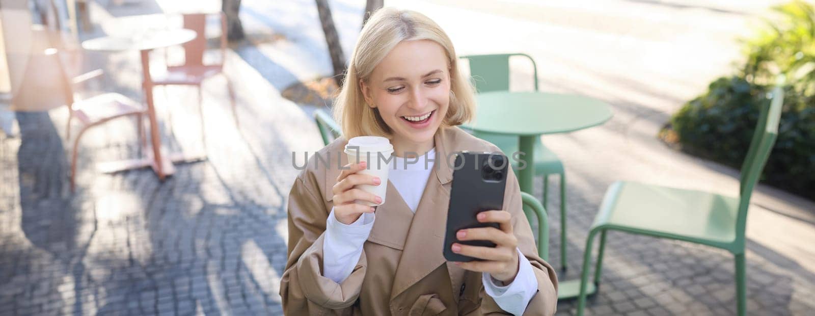 Portrait of cheerful, beautiful young woman taking selfie, chatting on video using smartphone, posing outdoors in coffee shop.