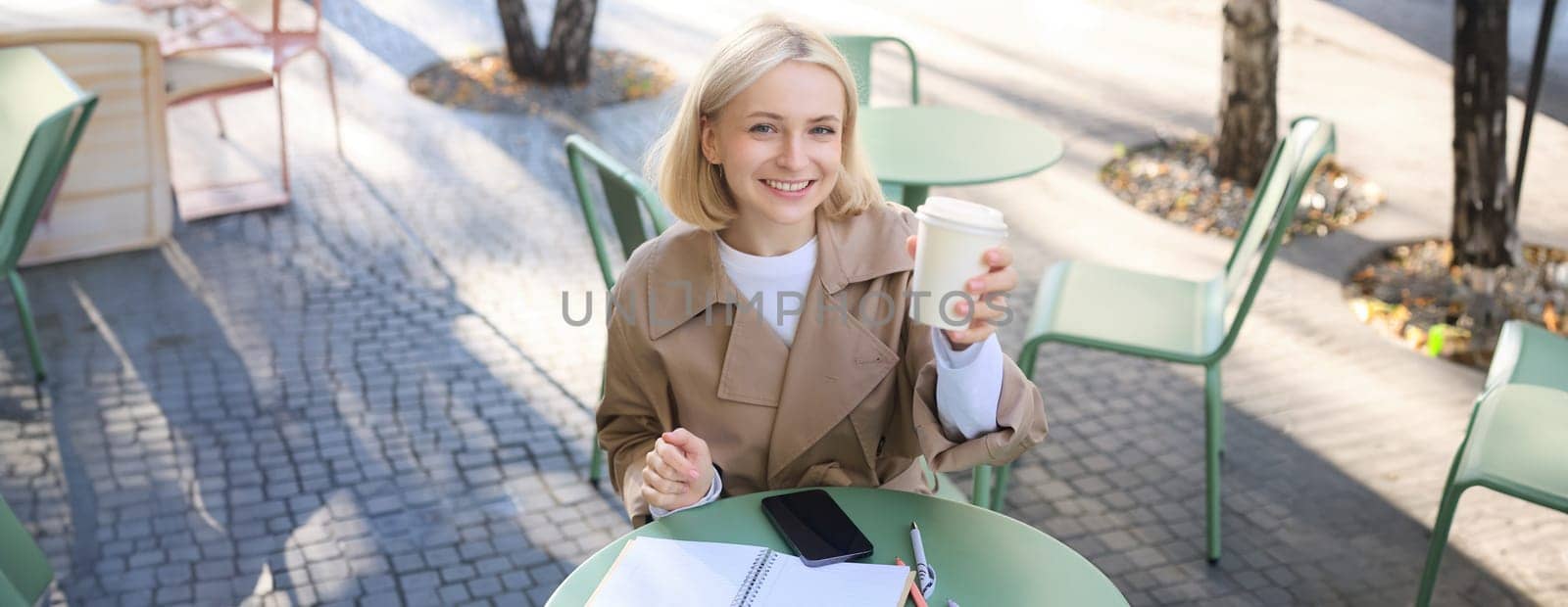 Beautiful blond woman, smiling, showing white takeaway coffee cup, drinking beverage in cafe shop, sitting outdoors by Benzoix