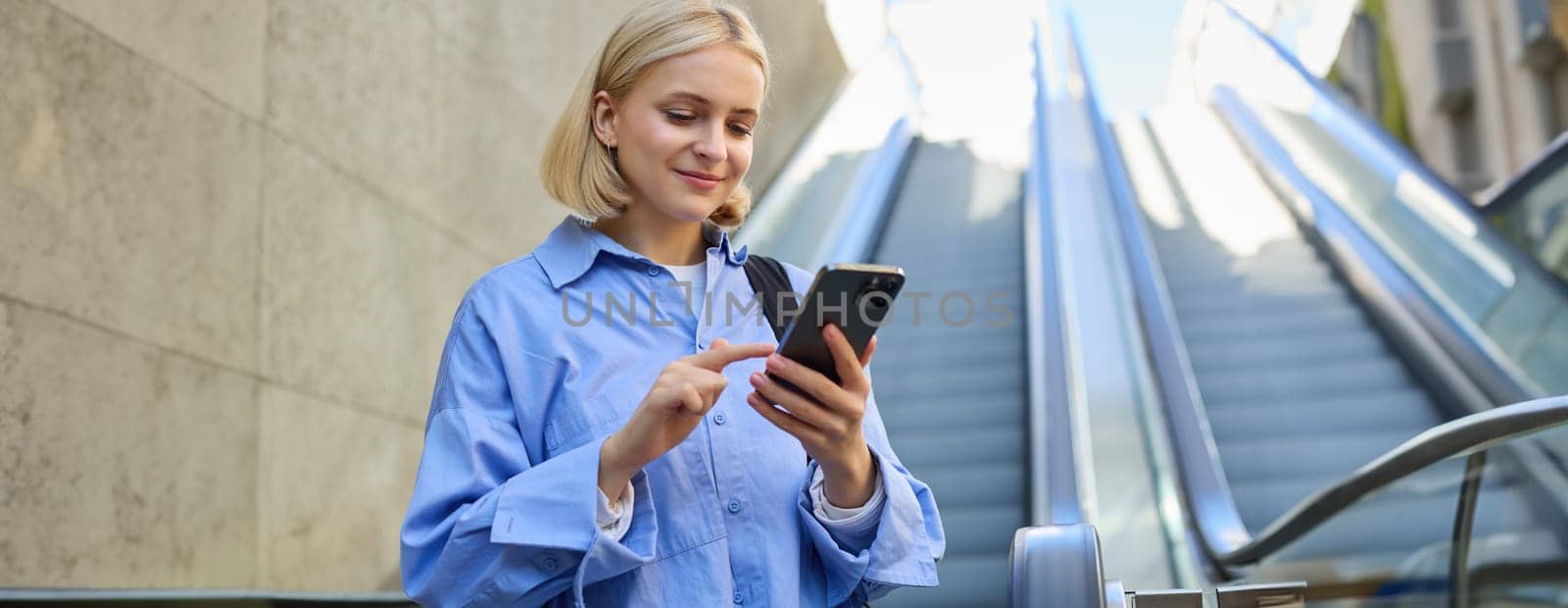 Portrait of young stylish woman, female employee, standing near escalator, using mobile phone, sending message on smartphone, tap screen, smiling and reading on telephone.