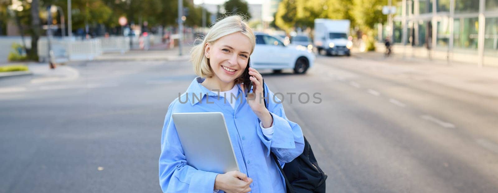 Portrait of young modern female model in blue shirt, talking on mobile phone, chatting with someone while walking along the street, holding laptop by Benzoix