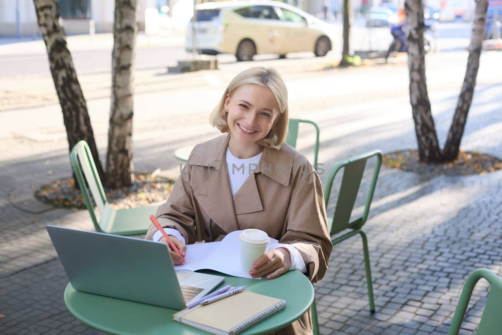 Image of young blond modern woman, sitting with laptop outside in cafe, drinking coffee drink and working on project, using computer.