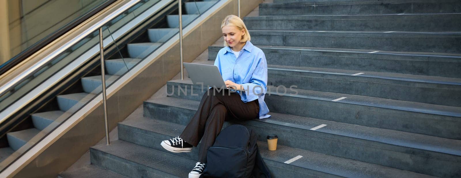 Freelance employee, woman working on her project, using laptop, sitting outdoors on stairs, drinking takeaway coffee by Benzoix