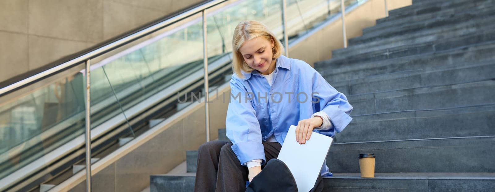 Portrait of young modern woman, student freelancer, sitting on public stairs outdoors, putting away her laptop, packing backpack, drinking coffee outside.