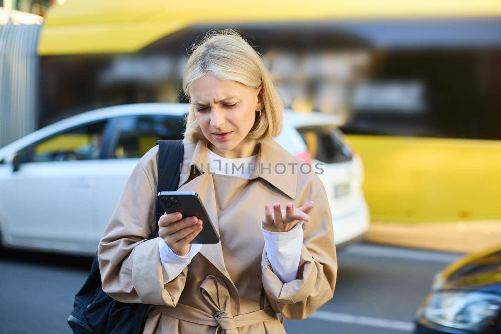Portrait of young confused blonde woman, standing on busy street with cars behind her, looking at smartphone with puzzled face, shrugging shoulders, reading smth with clueless expression.