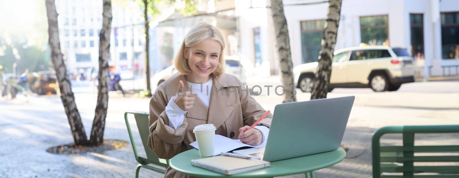 Portrait of beautiful blond female model, showing thumbs up, sitting with laptop and study material in coffee shop, gives approval. Lifestyle concept