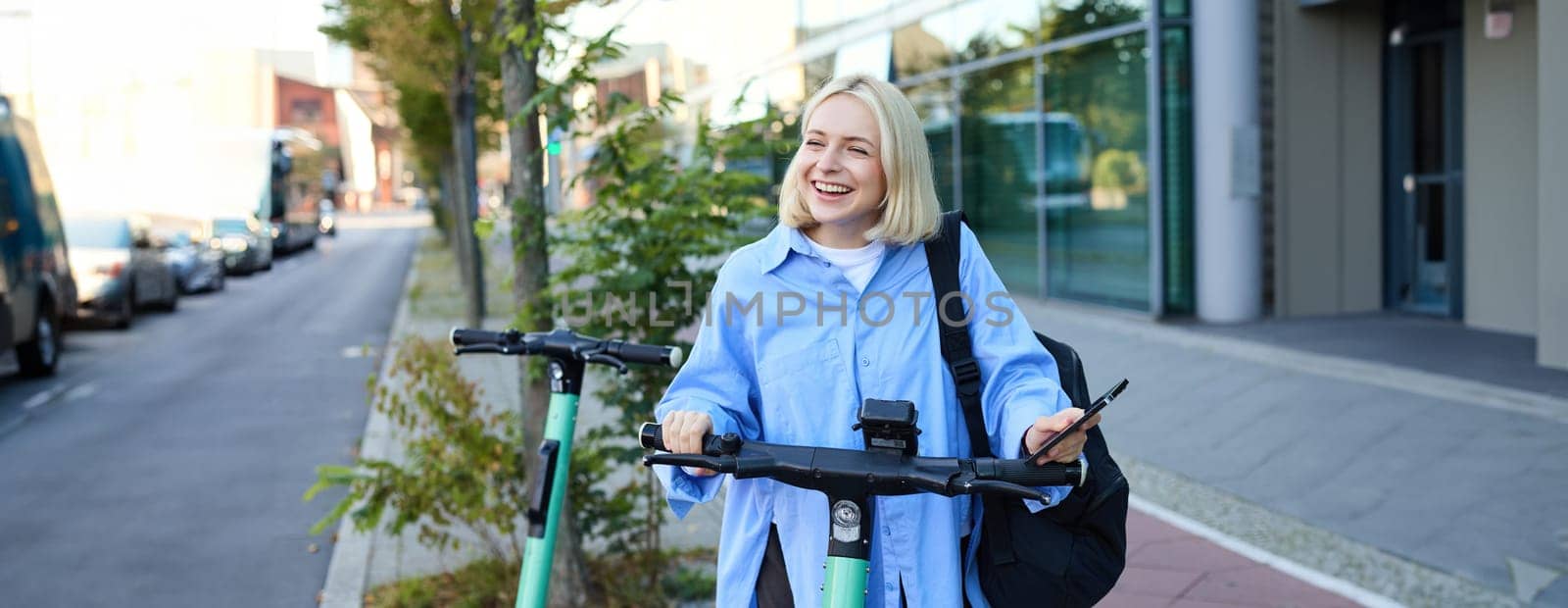 Portrait of smiling blond woman, college student using smartphone app to scan QR code on green electric scooter on street, renting a ride.