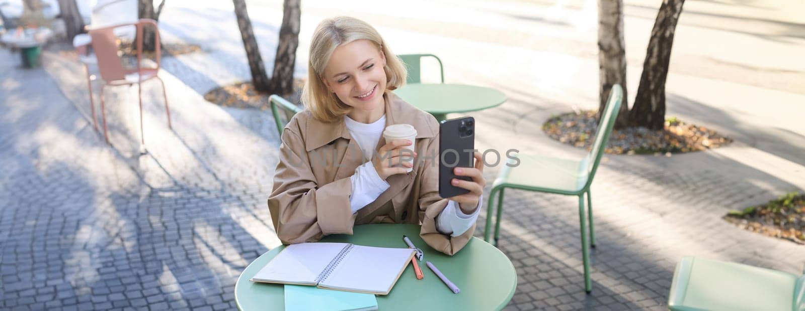 Image of stylish young woman, student taking selfie in cafe on street, posing with her favourite drink, enjoying coffee and making content for social media blog.