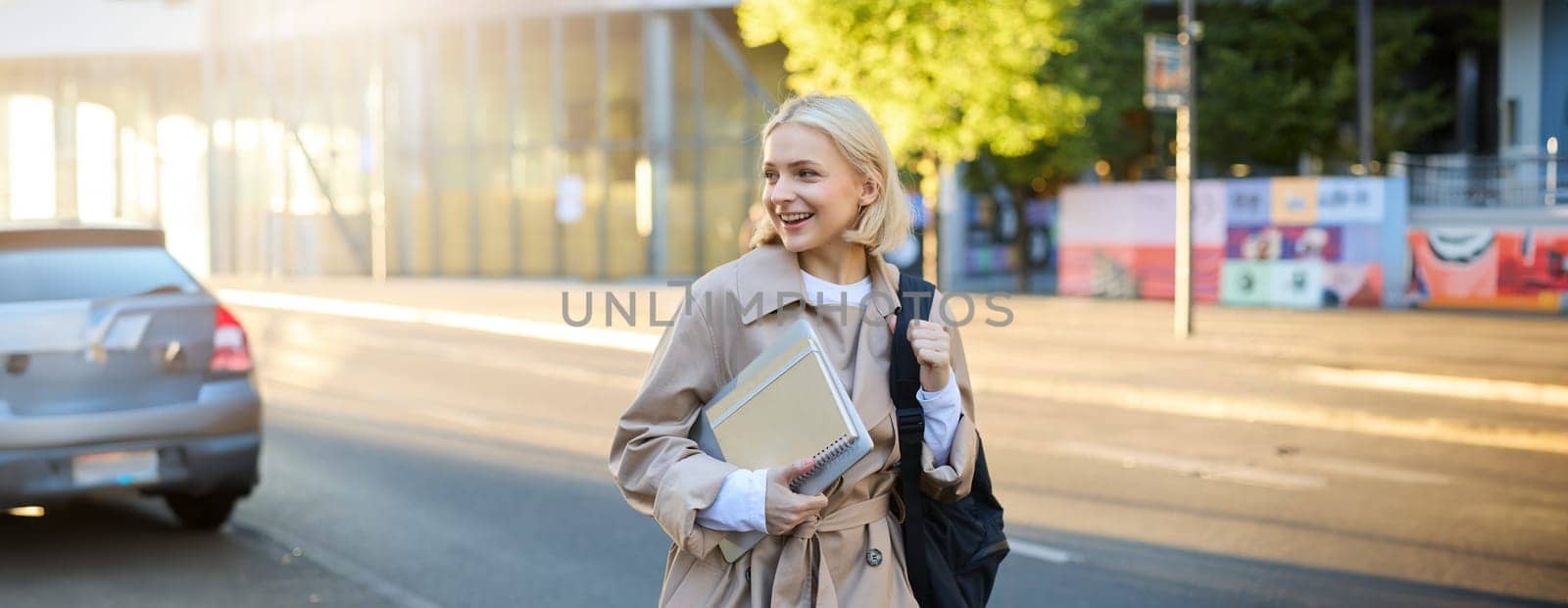 Lifestyle portrait of young woman walking on street with backpack and notebook, looking aside, saying hello to someone on her way to university or college.