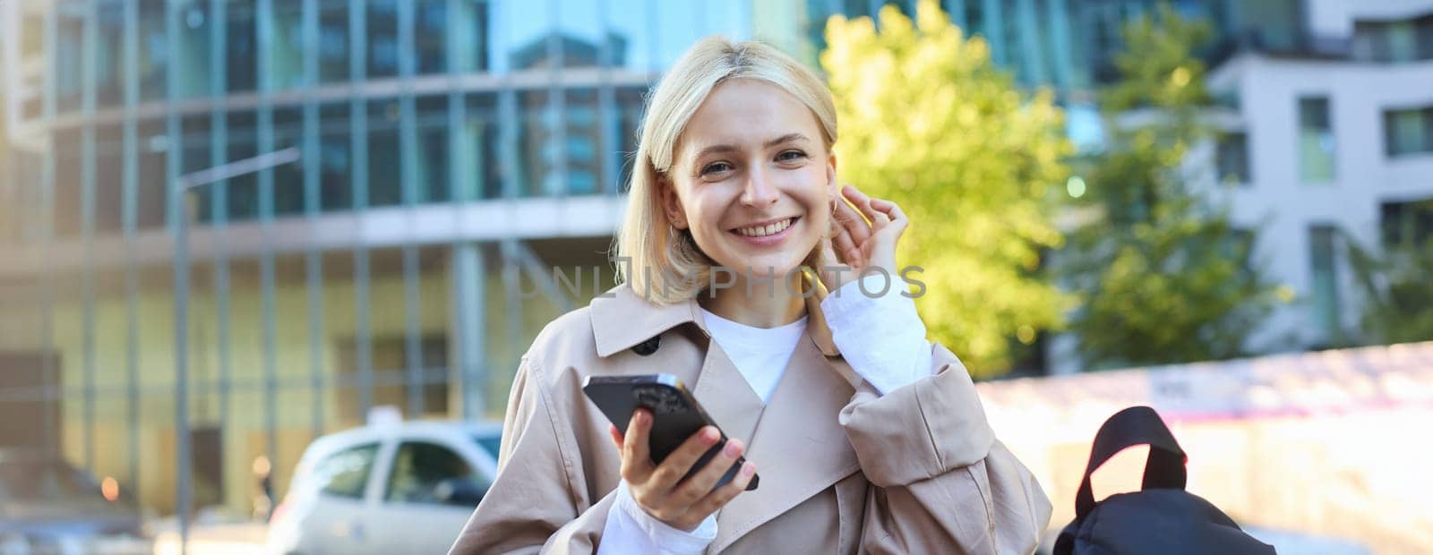 Portrait of carefree smiling woman on street, holding mobile phone, tuck hair behind ear and looking happy at camera, waiting for someone outside.
