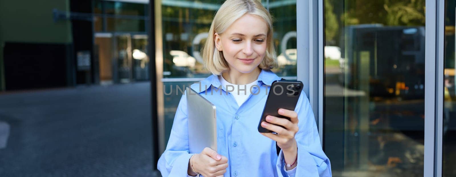 Lifestyle shot of young smiling woman, student or employee near office building, holding mobile phone and laptop in hands, looking happy, checking notification on smartphone by Benzoix